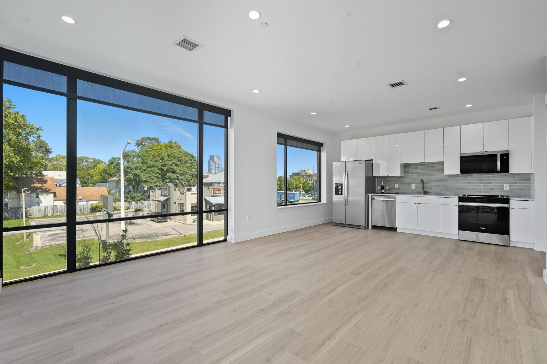 Bright modern apartment with large windows, white kitchen, stainless steel appliances, and urban view.