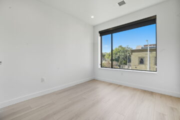 Bright modern bedroom with large window overlooking neighborhood, minimalistic design, clean white walls, and light wooden flooring.