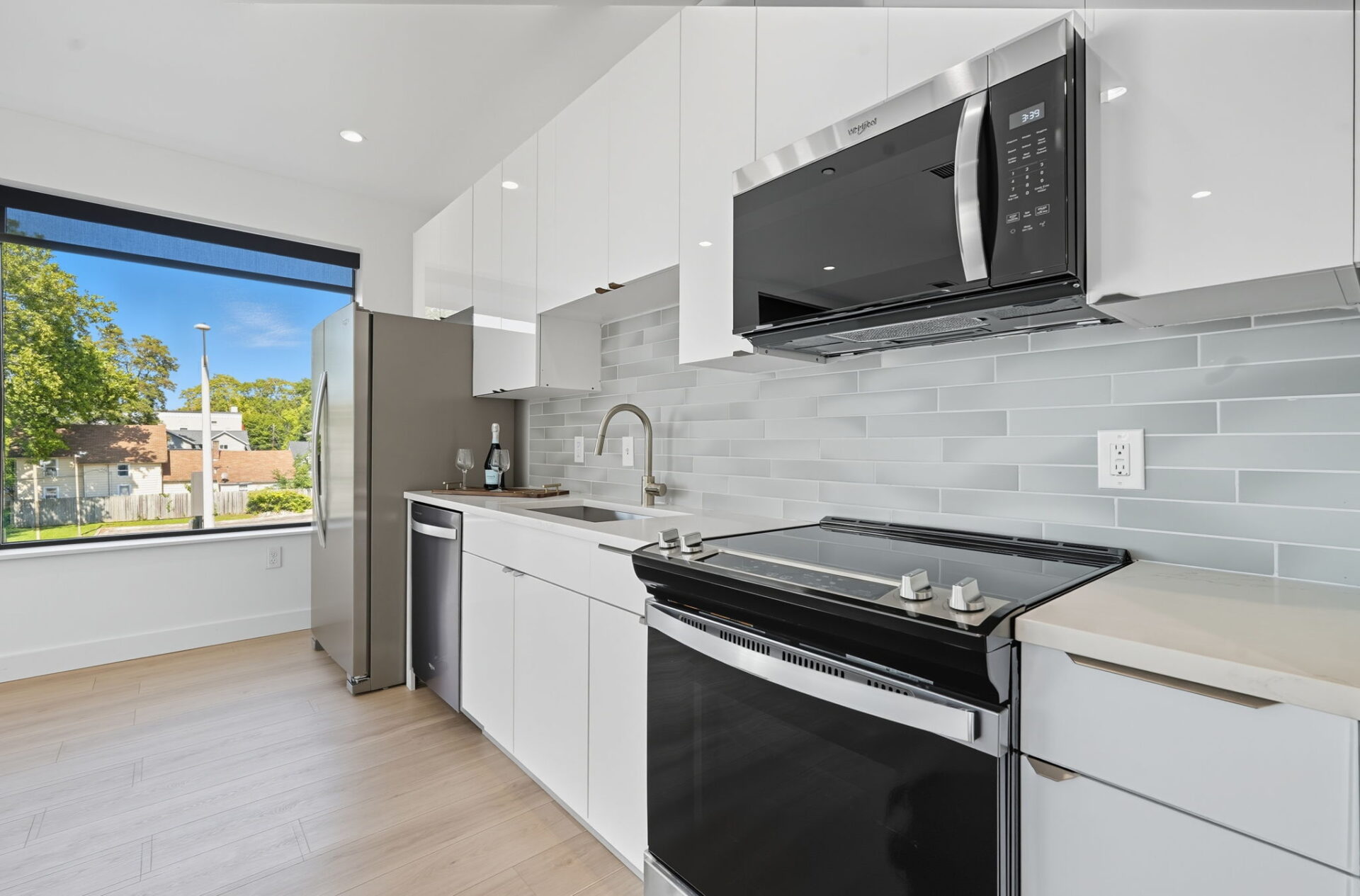 Modern white kitchen with stainless steel appliances and large window showcasing outdoor greenery.