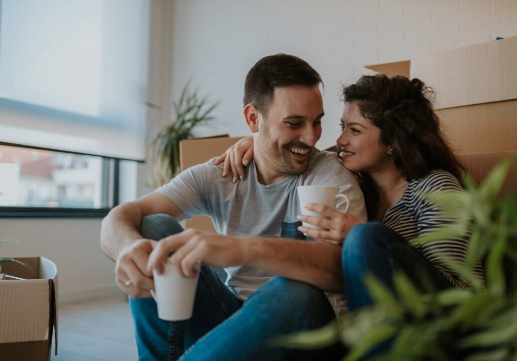 Relaxing in new house. Cheerful young couple sitting on the floor and drinking coffee while cardboard boxes laying all around them.