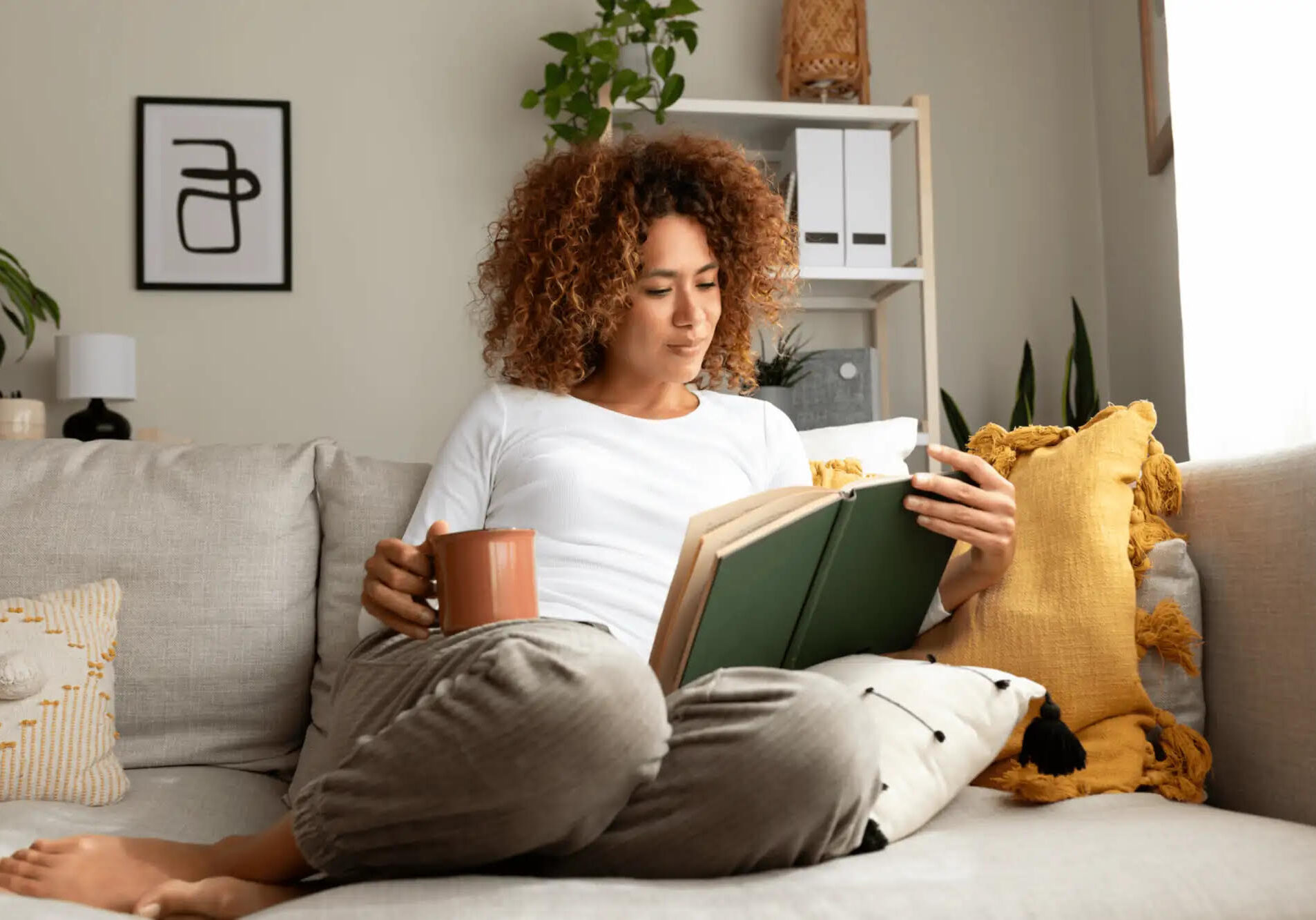 Woman with curly hair reading book on couch with coffee